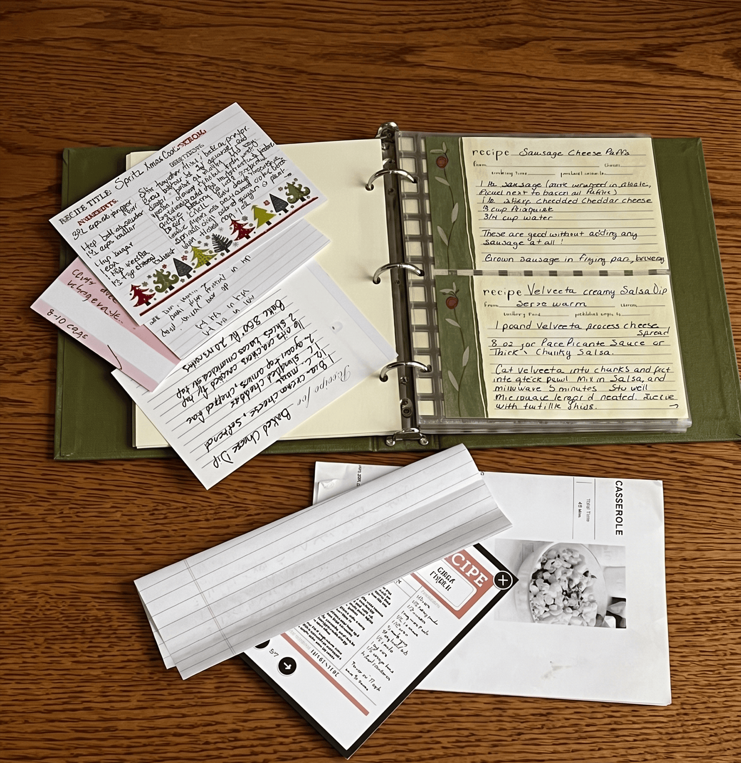 A handwritten family recipe book opened to two recipes, surrounded by scattered note cards, printed recipes, and handwritten notes on a wooden table.